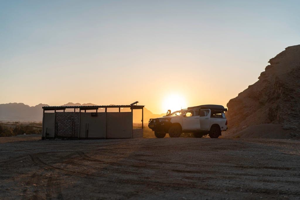 Weißes Geländefahrzeug neben Schutzhütte in felsiger Wüstenlandschaft bei Sonnenuntergang.