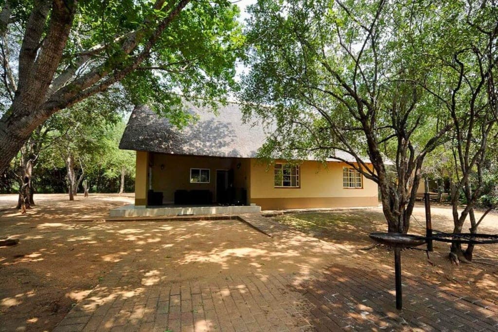 Einfache Lodge mit Strohdach und Veranda in schattigem Baumbestand, Sandboden und gepflastertem Weg