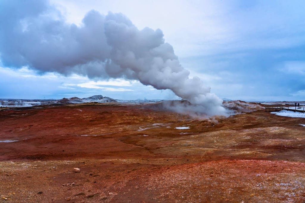 Geothermisches Feld mit rotem Boden und dichter Dampfwolke unter bewölktem Himmel