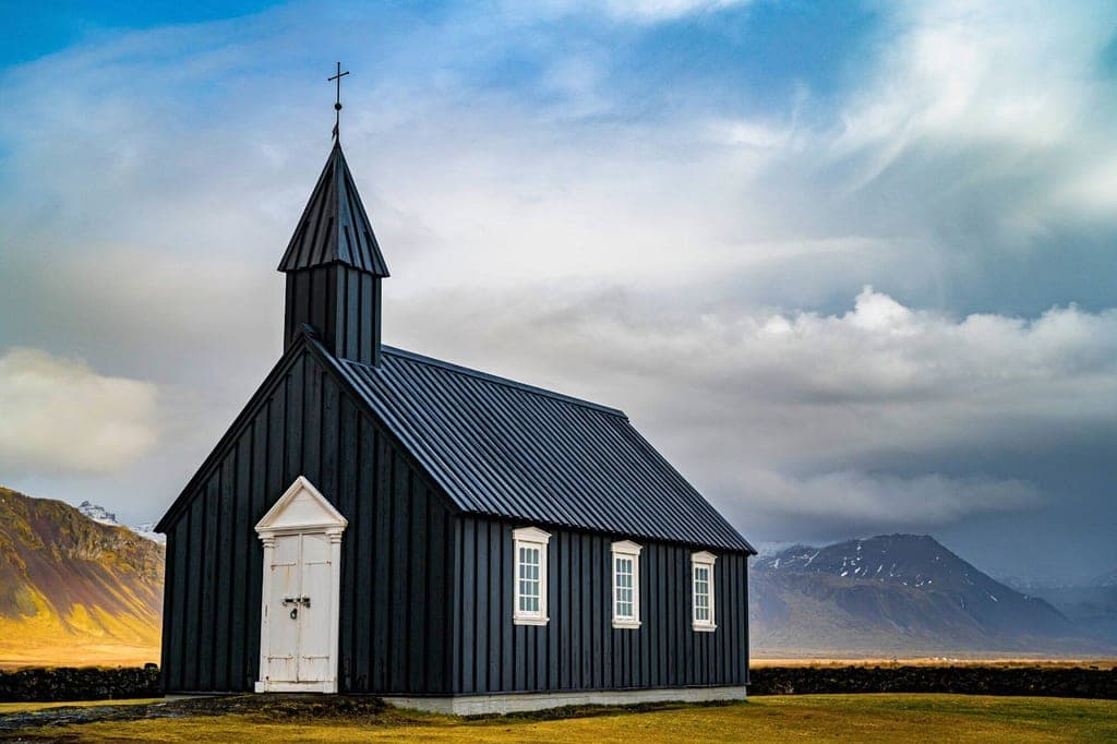 Schwarze Holzkirche vor Bergen und dramatischem Wolkenhimmel in karger Landschaft.