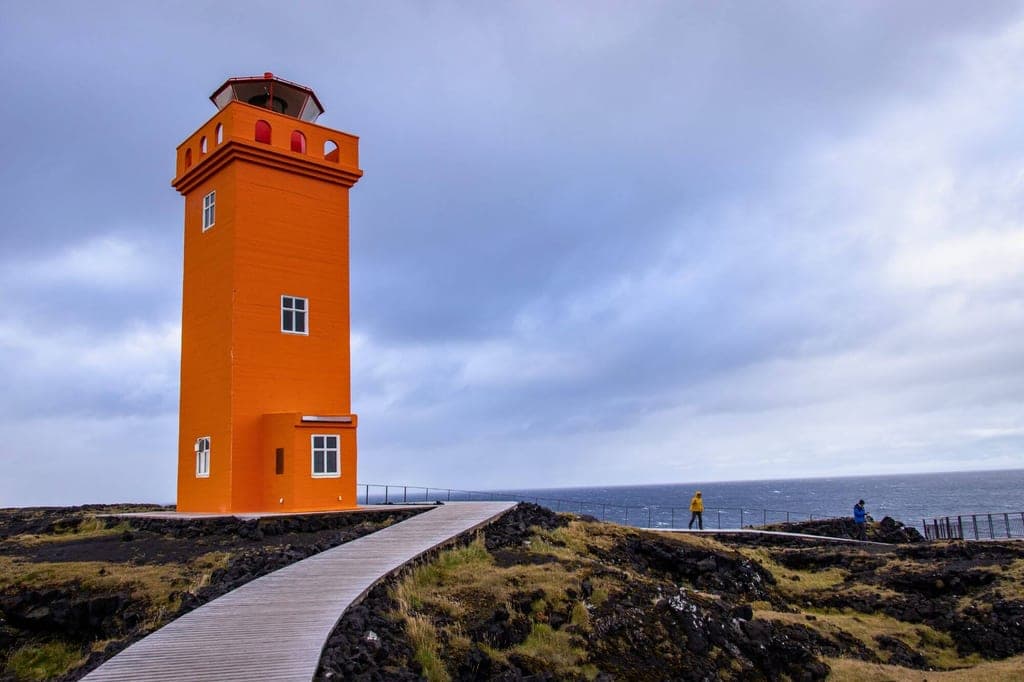 Oranger Leuchtturm an felsiger Küste mit Holzsteg, Meer und bewölktem Himmel, zwei Personen am Geländer.