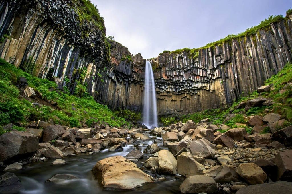 Wasserfall fällt zwischen hohen Basaltsäulen in eine felsige Schlucht, Bach im Vordergrund.