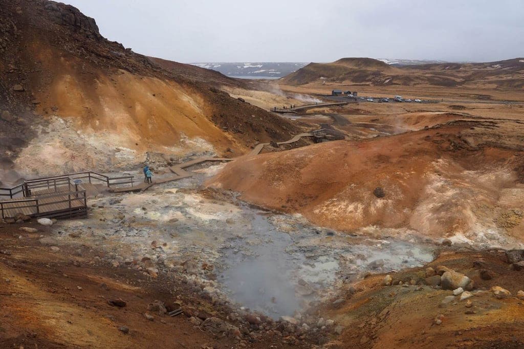 Geothermisches, dampfendes Gebiet mit bunten Hügeln und Holzstegen bei bewölktem Himmel.