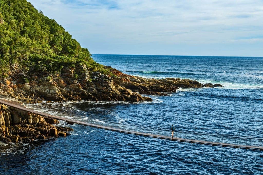 Person auf Hängebrücke über rauem Meer vor bewaldeter Felsenküste im Tsitsikamma National Park, Südafrika.