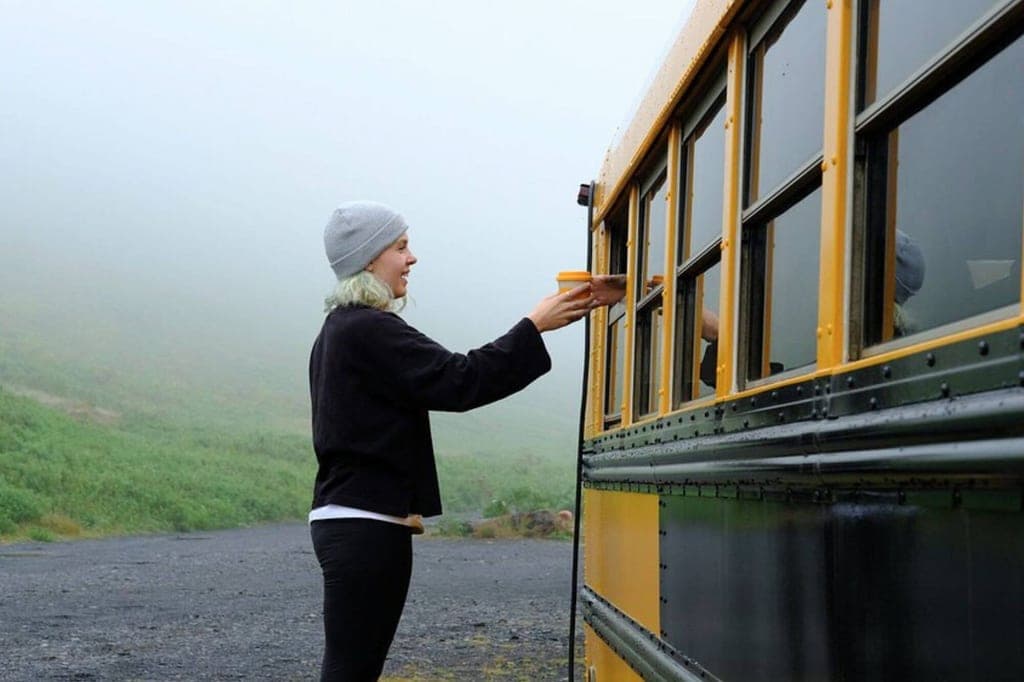 Person reicht Becher durch geöffnetes Fenster an einen gelben Bus bei nebliger Landschaft.