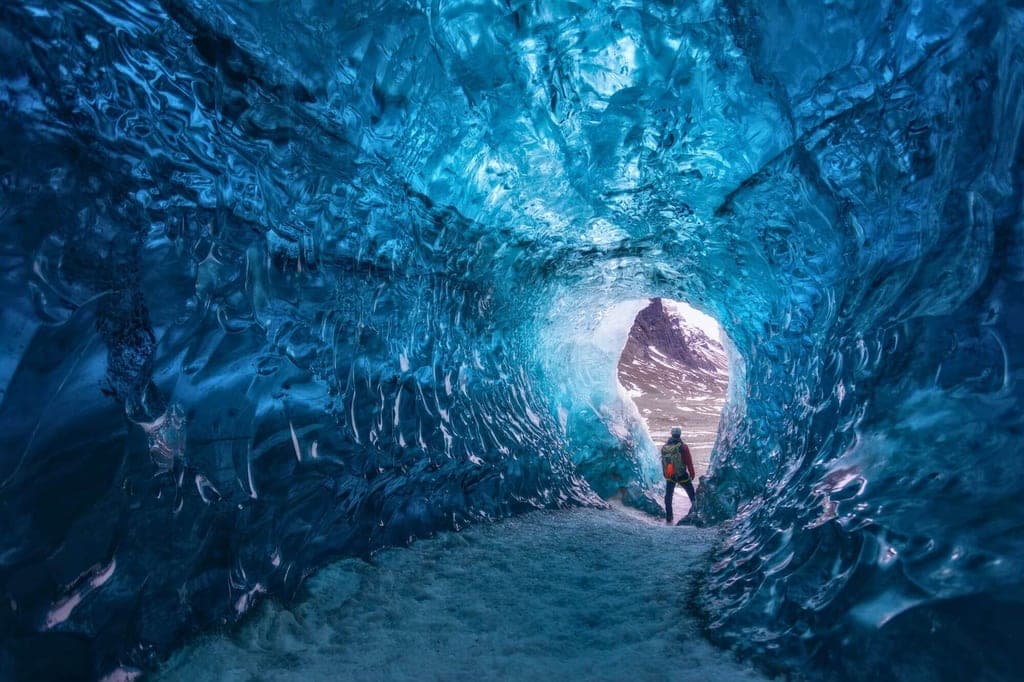 Blick aus einer blauen Eishöhle auf eine Person am Höhlenausgang bei Tageslicht.