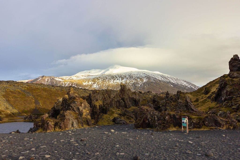 Vulkanlandschaft mit schwarzem Kies, zerklüfteten Felsen und schneebedecktem Berg unter bewölktem Himmel.