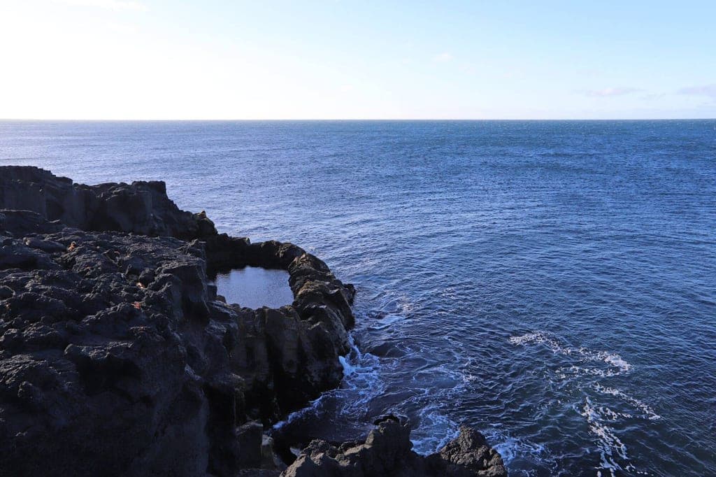 Dunkle Lavafelsen und Gezeitentümpel an einer ruhigen Küste mit weitem, blauem Meer bis zum Horizont.