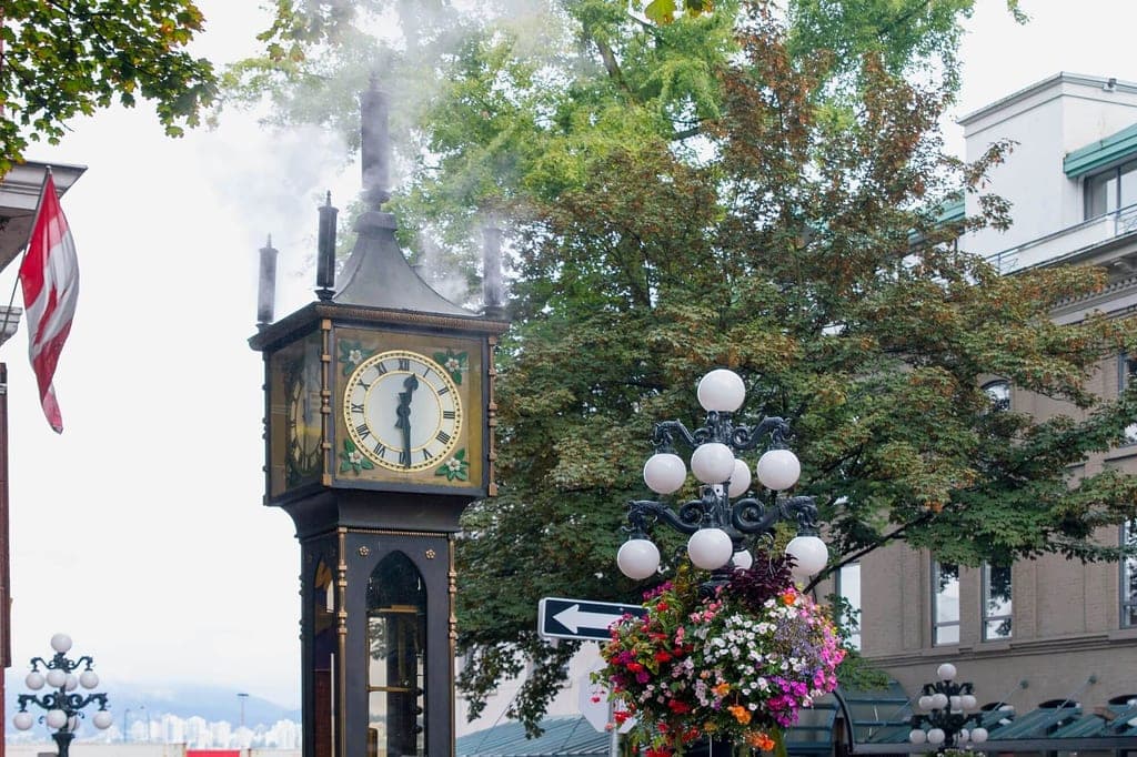 Dampfstanduhr mit aufsteigendem Dampf neben einer Straßenlaterne mit Blumenampel