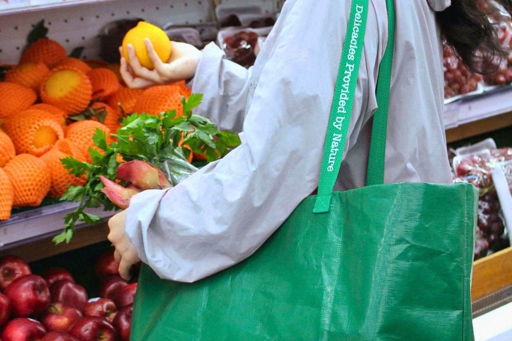 Seitenaufnahme einer einkaufenden Person mit grüner Stofftasche, hält eine Orange vor Obstregalen.