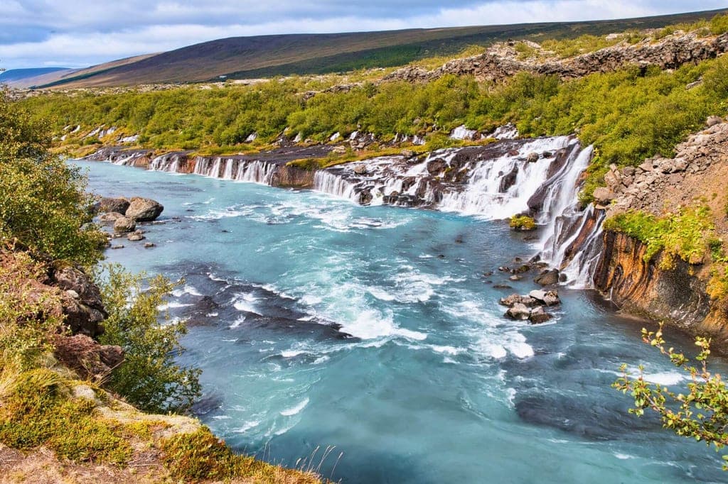 Mehrere Kaskaden eines Wasserfalls in einen türkisfarbenen Fluss, grünes Ufer und bewachsene Hügel im Hintergrund.