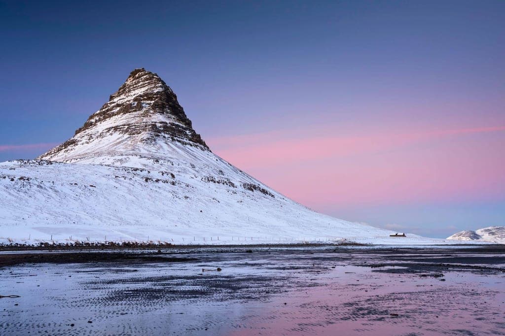 Schneebedeckter markanter Berg an der Küste mit pastellfarbenem Dämmerungshimmel und Spiegelung im nassen Sand.