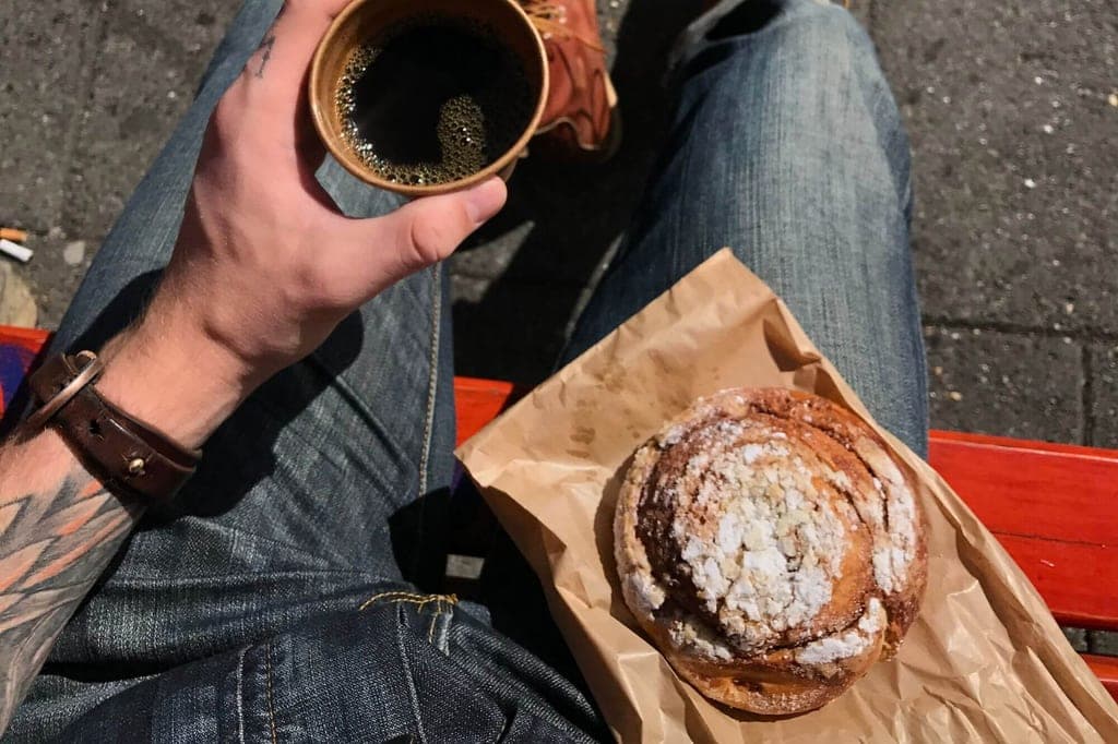 Ich-Perspektive: tätowierter Arm mit Kaffee und Zimtschnecke auf Jeans vor roter Bank