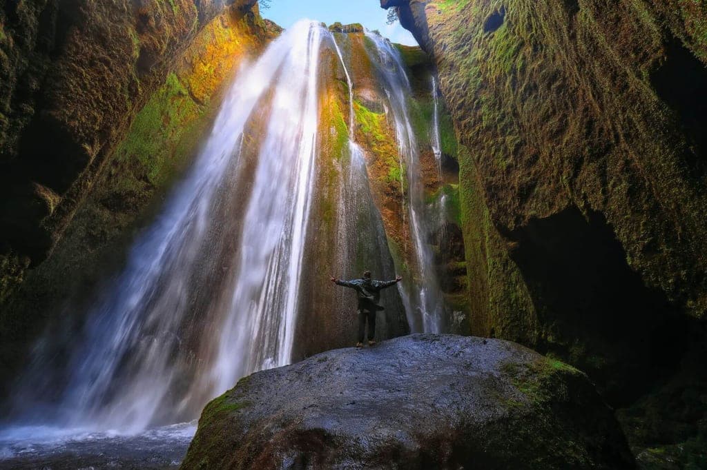 Person mit ausgebreiteten Armen auf Felsen vor moosbedecktem Wasserfall in schmaler Schlucht