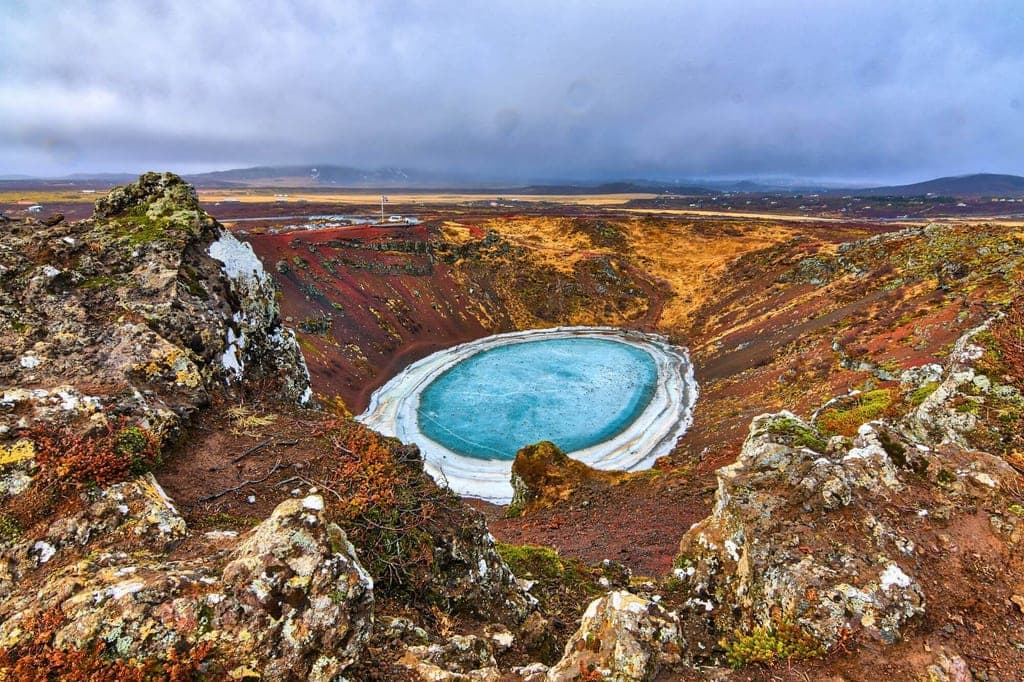 Blick von einem felsigen Kraterrand auf einen kreisrunden, teilweise gefrorenen Kratersee mit türkisblauem Wasser.