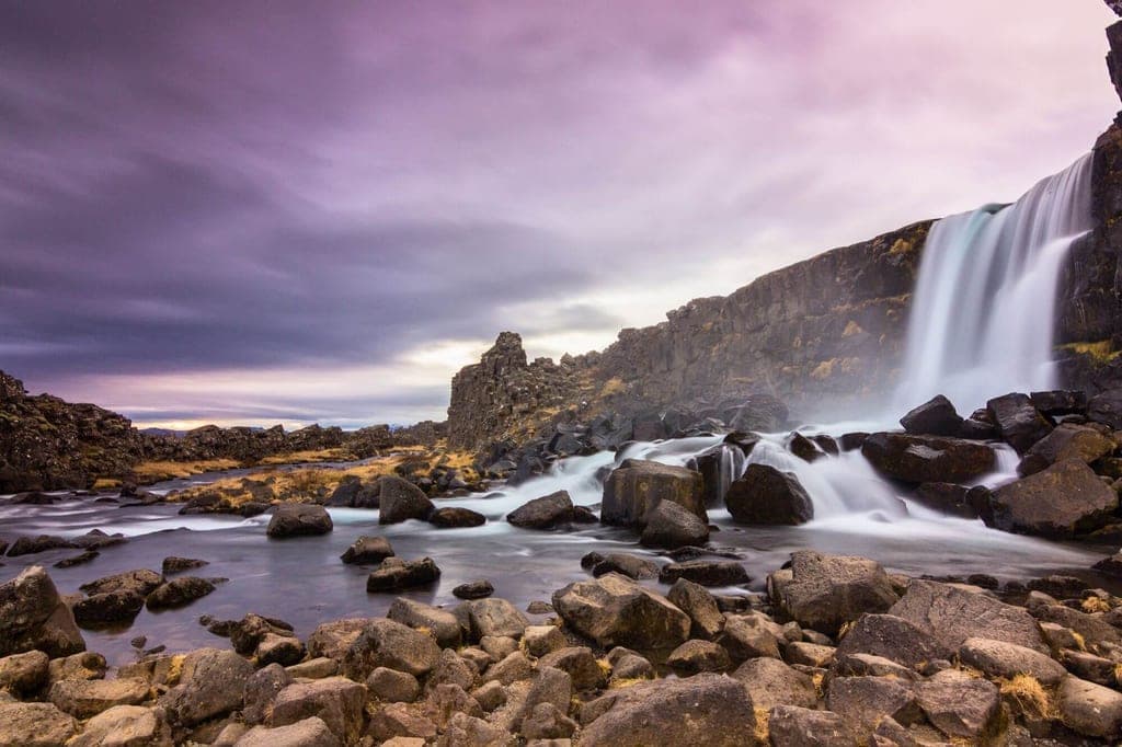 Wasserfall stürzt über dunkle Felsen in einen von Steinen durchsetzten Fluss bei dämmerndem violettem Himmel.