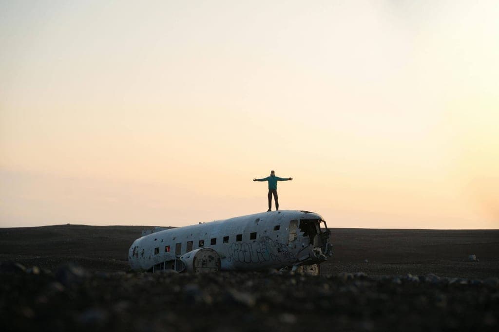 Person steht mit ausgestreckten Armen auf dem Flugzeugwrack in weiter Landschaft bei Sonnenuntergang.