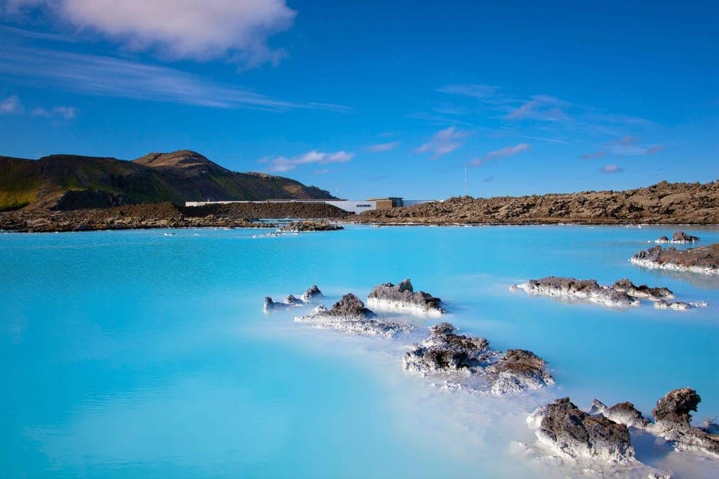 Milchblaues Thermalbecken mit Lavafelsen und Hügeln unter klarem blauem Himmel.