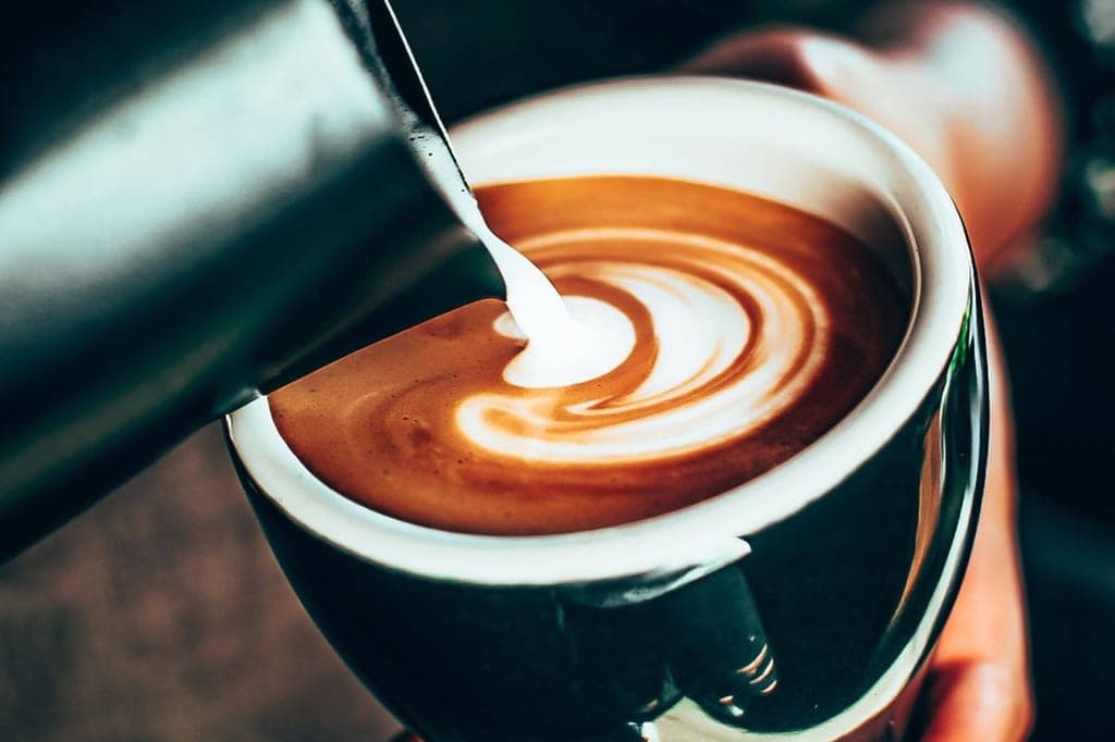 Nahaufnahme: Milch wird in eine Tasse Kaffee gegossen, sichtbare Latte-Art auf der Oberfläche.