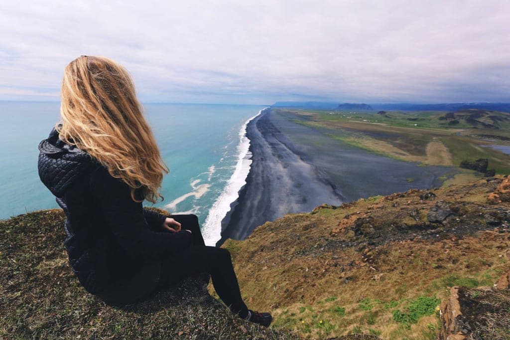 Person sitzt auf Klippe und blickt über langen schwarzen Sandstrand und ruhige See unter Wolken