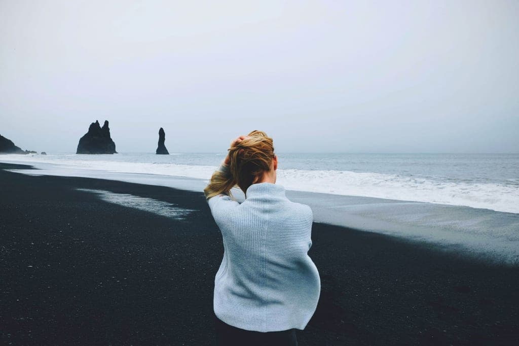 Person von hinten am schwarzen Sandstrand vor felsigen Seeformationen bei bewölktem Himmel.