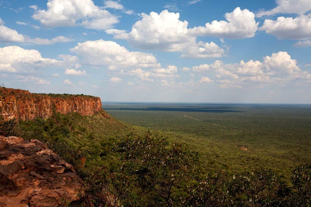 Blick von einem Felsplateau über weite Savannenebene mit roter Klippe und Cumuluswolken