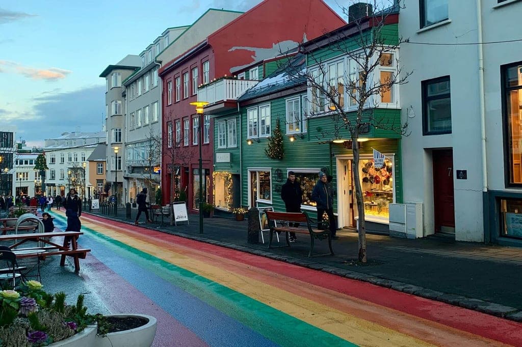 Straße mit in Regenbogenfarben gestrichenem Gehweg vor bunten Häusern und beleuchteten Schaufenstern