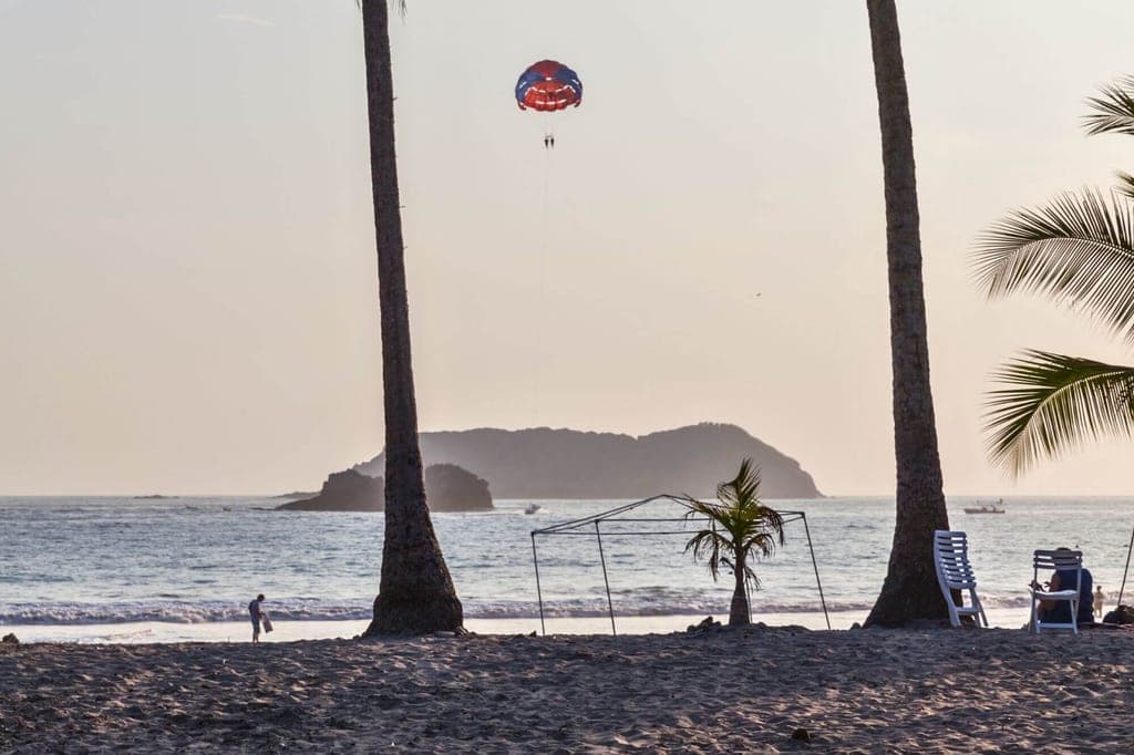 Strand bei Abendlicht mit Palmen, Liegestühlen und einem Parasail über dem Meer