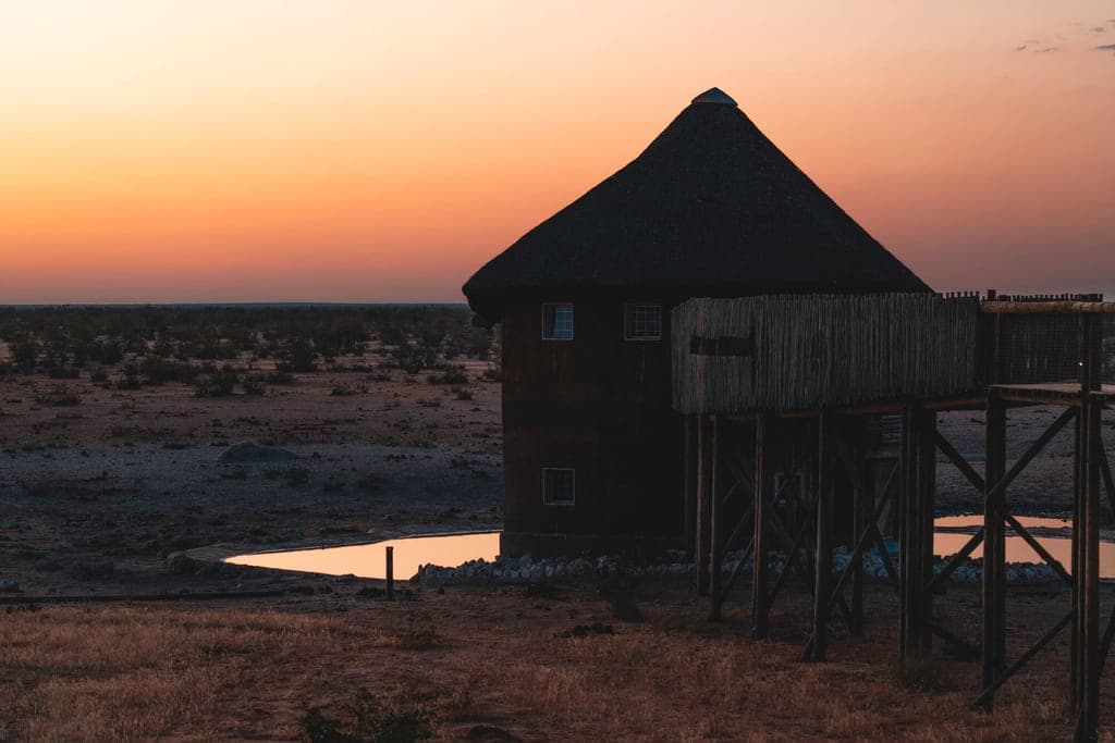 Strohgedeckte Beobachtungshütte auf Pfählen neben einem Wasserloch bei Sonnenuntergang