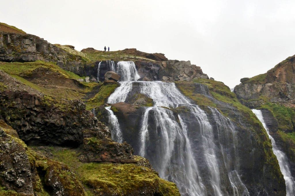 Mehrstufiger Wasserfall über moosige Felsen unter bedecktem Himmel, zwei Personen auf dem Plateau.