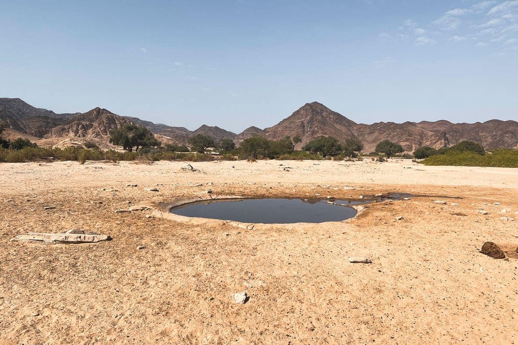 Trockene Wüstenlandschaft mit kleinem Wasserloch und karger Bergkulisse bei klarem Himmel.