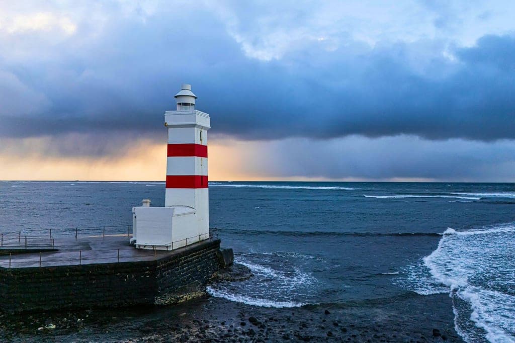 Weißer Leuchtturm mit roten Ringen an einer Mole vor stürmischem Meer und gelbem Licht am Horizont.