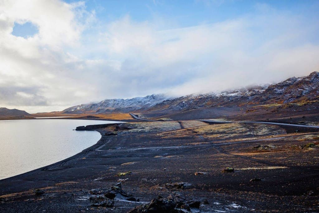 Weite Vulkanlandschaft mit See, dunklem Kiesstrand, Straße und schneebedeckten Hügeln unter bewölktem Himmel.