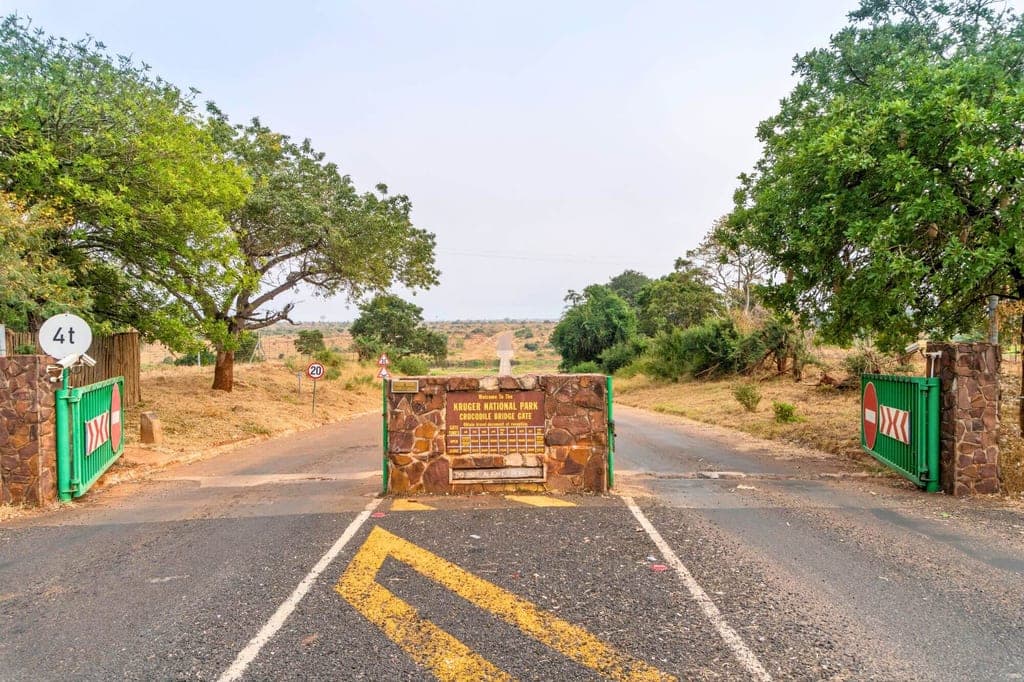 Frontansicht eines Parkeingangs mit Steinbarriere und grünen Toren vor trockener Savannenlandschaft