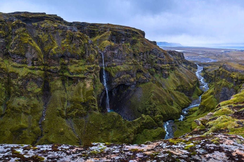 Blick von einem Felsrand in eine moosbedeckte Schlucht mit schmalem Fluss und hohem Wasserfall bei bewölktem Himmel.