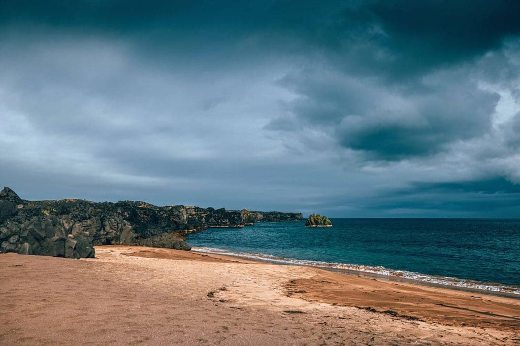 Sandstrand mit dunklen Felsklippen, einsamer Felsen im Meer und bewölktem Himmel