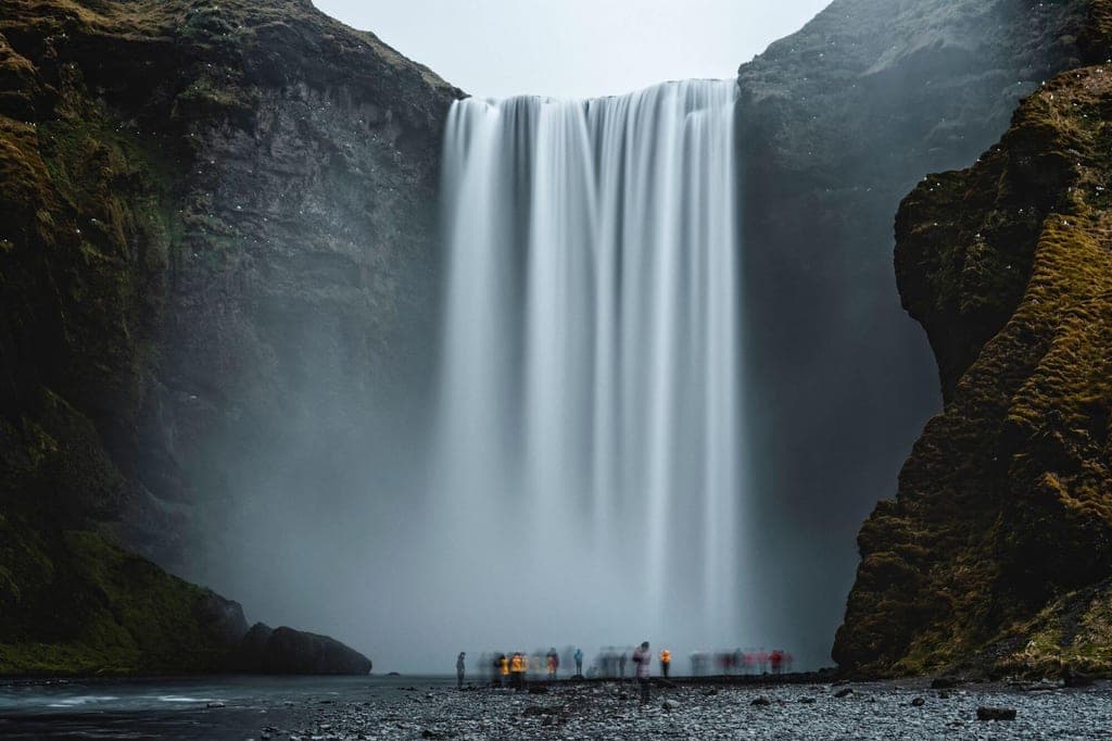 Breiter Wasserfall fällt von einer steilen Felswand, Menschen stehen unscharf am nebligen Ufer.