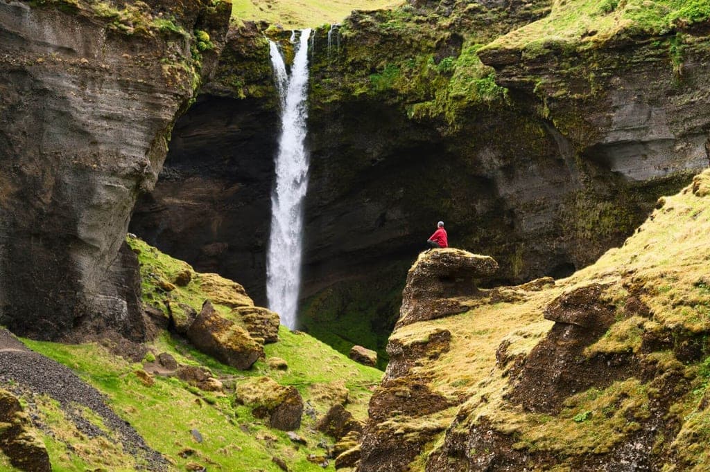 Weitaufnahme eines Wasserfalls in moosiger Schlucht; Person in roter Jacke sitzt auf einem Felsvorsprung.