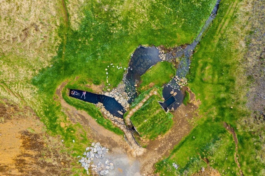 Vogelperspektive auf grasige Landschaft mit Bach, Felsbecken und zwei Personen im Wasser.